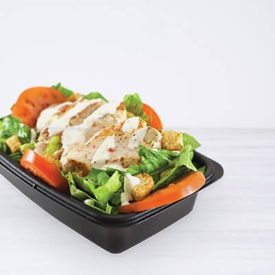 Salade dans un contenant noir, avec laitue, tomates, oignons, croutons, poulet et sauce ranch, sur fond blanc